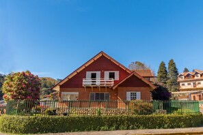 Uma casa vermelha com varanda, cercada por um muro e um viveiro verdes.