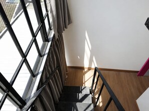 Una escala amb baranes metàl·liques i un paviment de fusta que condueix a una habitació amb una paret blanca i una finestra.
