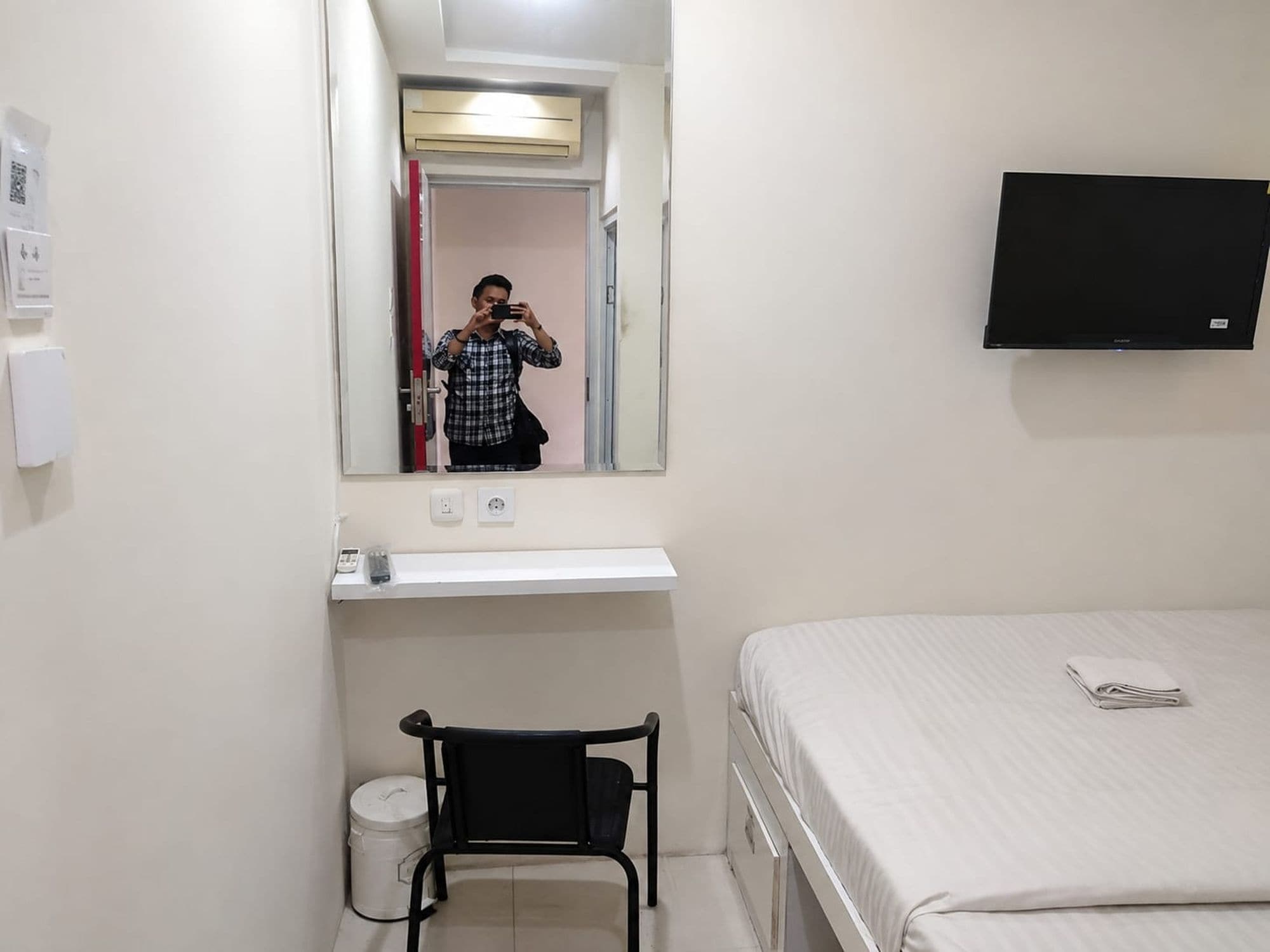 A hotel room with a bed, a chair, a mirror, a television, and a small shelf with a power outlet.