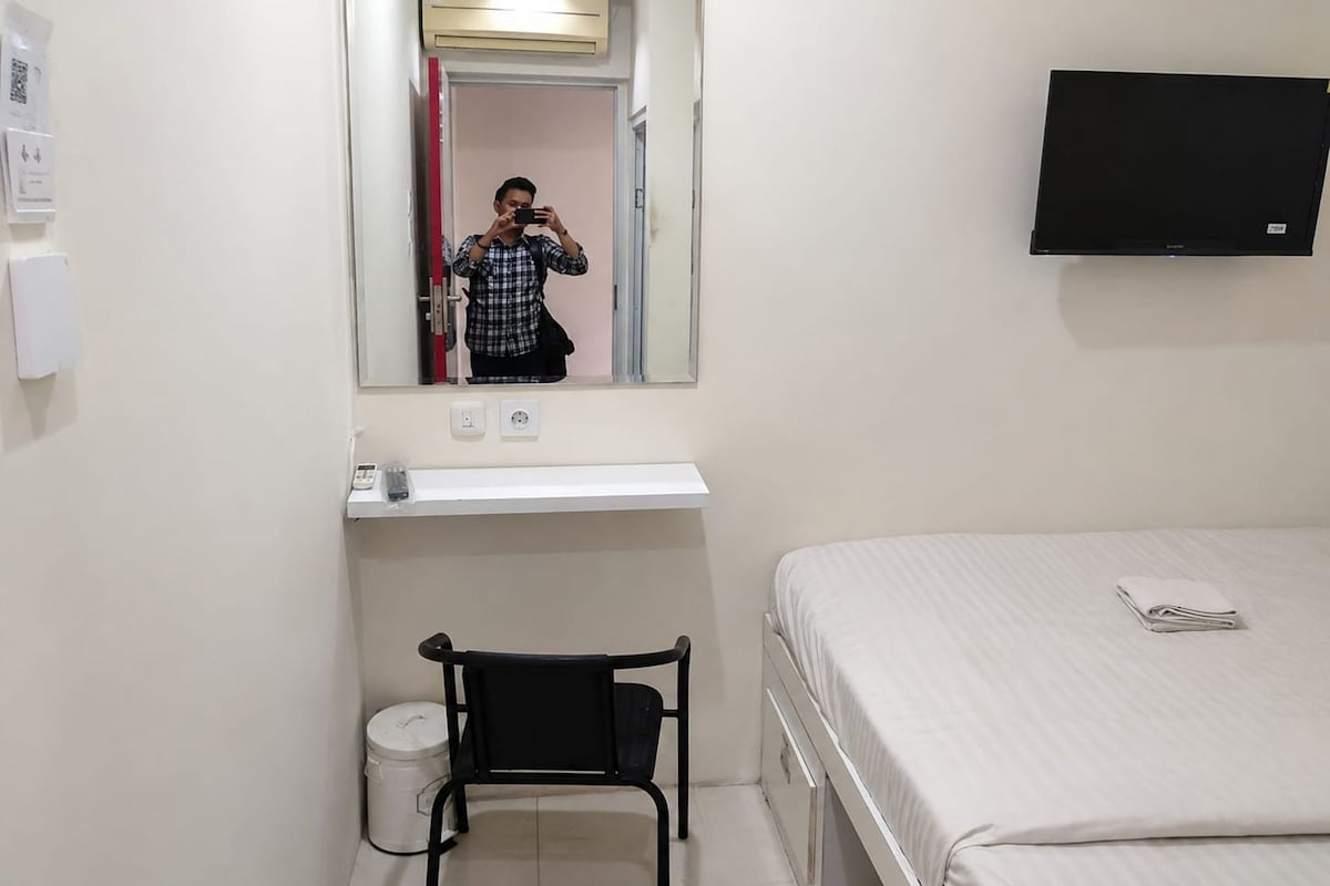 Habitació d'hotel amb un llit, una cadira, una taula petita, un mirall, un televisor i un safataire.