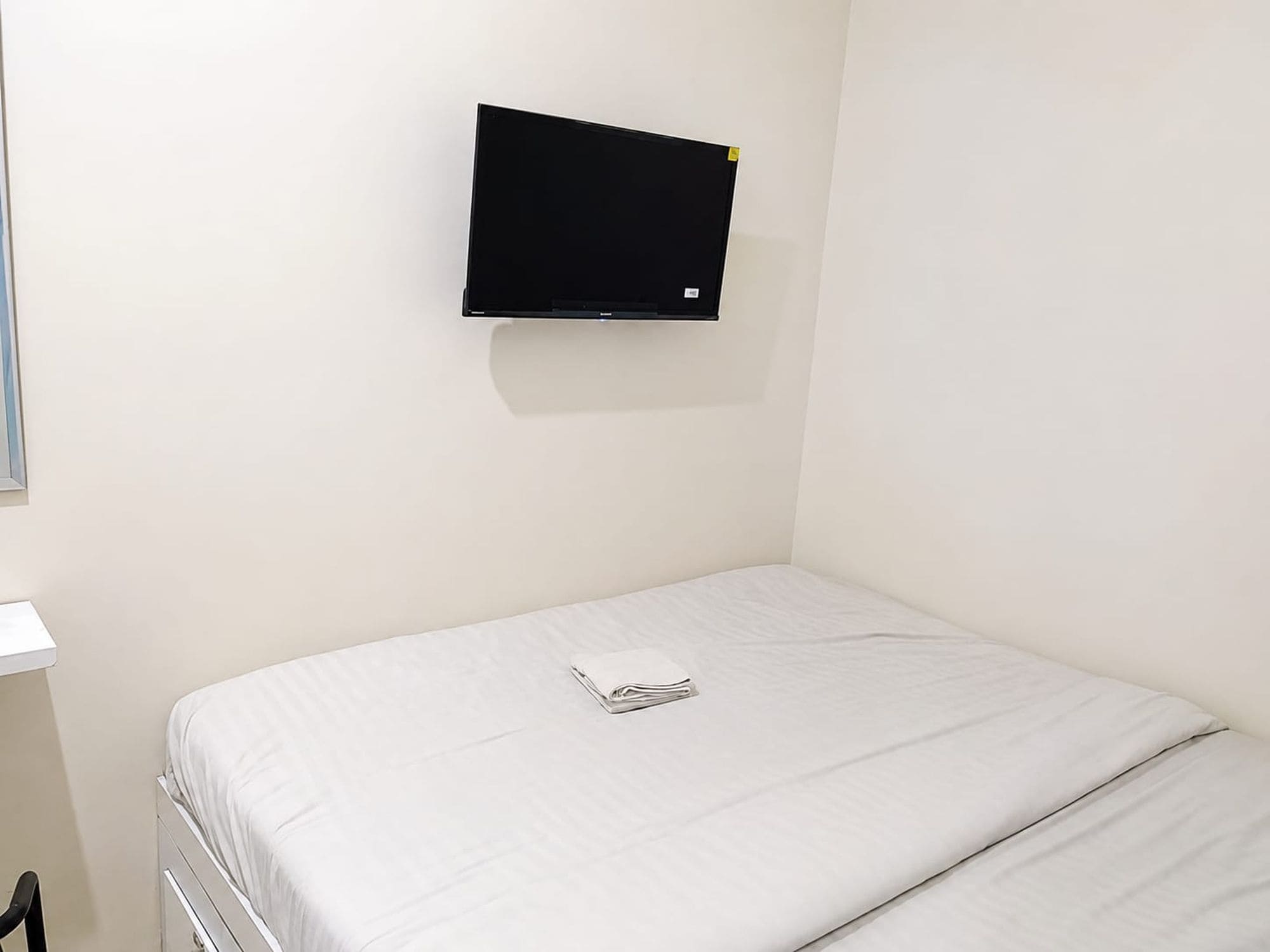 A single bed with a white mattress and a folded white towel. A black television is mounted on the wall above the bed.