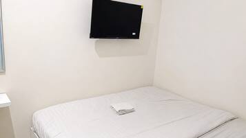 A single bed with a white mattress and a folded white towel. A black television is mounted on the wall above the bed.