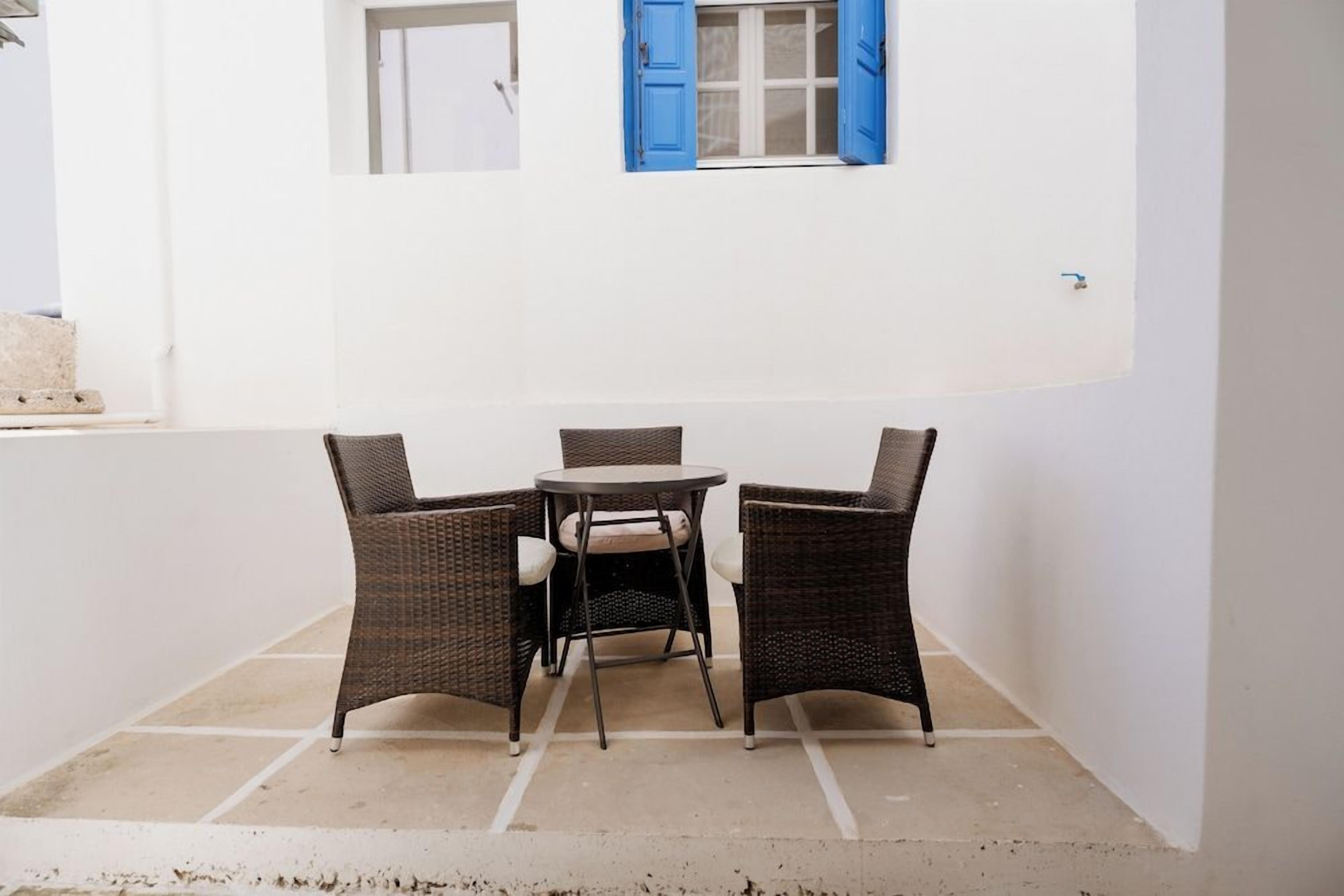 Un pequeño espacio exterior con sillas de mimbre y una mesa redonda, frente a una pared blanca con un contraventano azul.