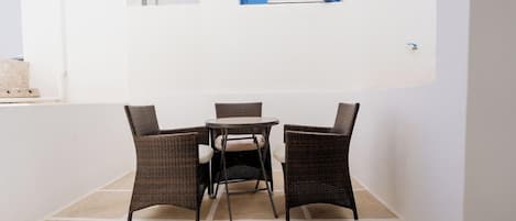 Un pequeño espacio exterior con sillas de mimbre y una mesa redonda, frente a una pared blanca con un contraventano azul.
