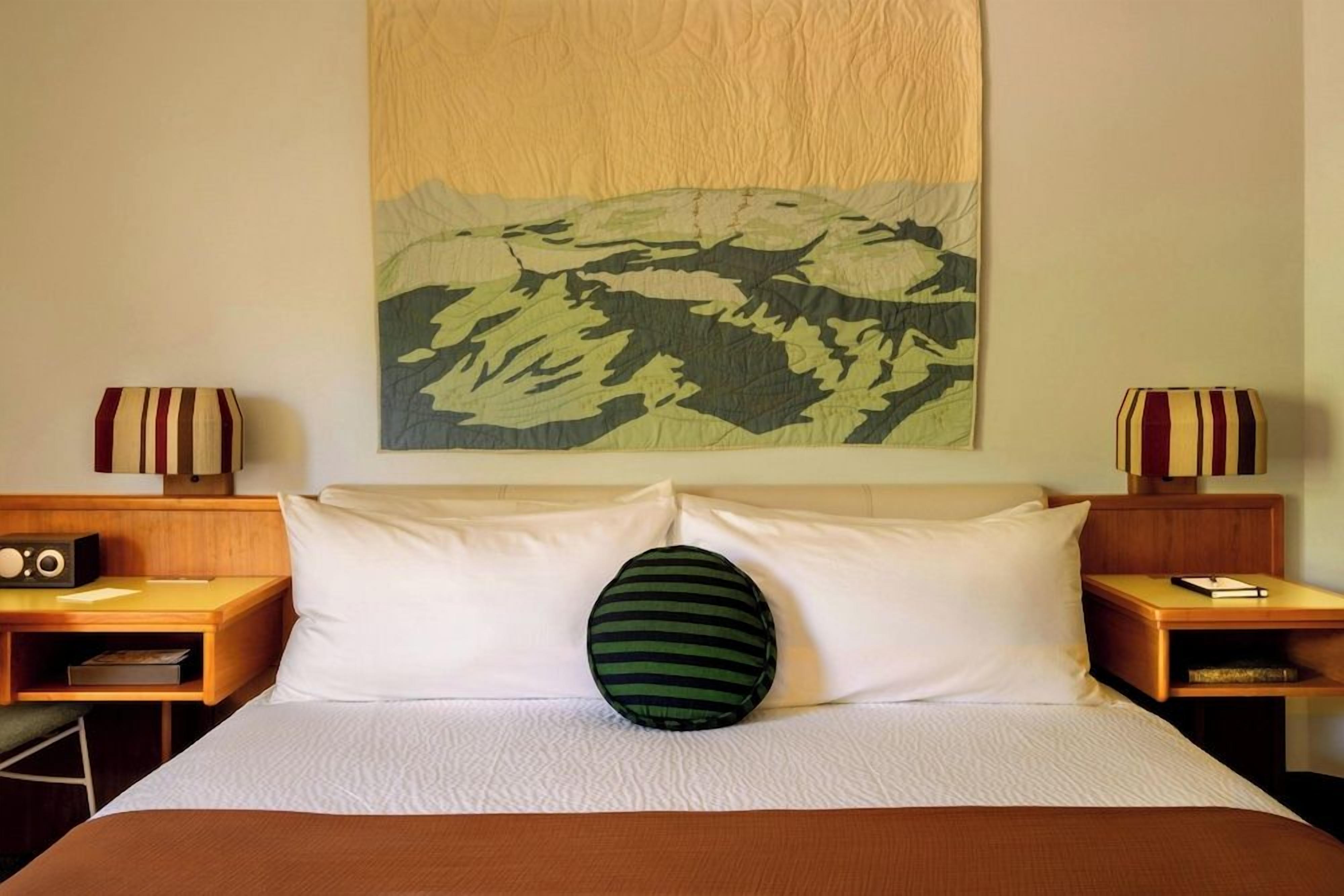 Habitación de hotel con cama, dos mesitas de noche, una radio, almohada y un cuadro colgado en la pared.
