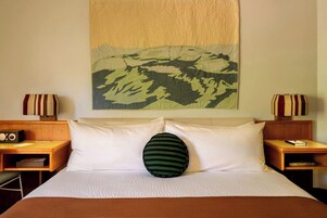 A hotel room with a bed, two bedside tables, a radio, a pillow, and a wall-mounted painting.