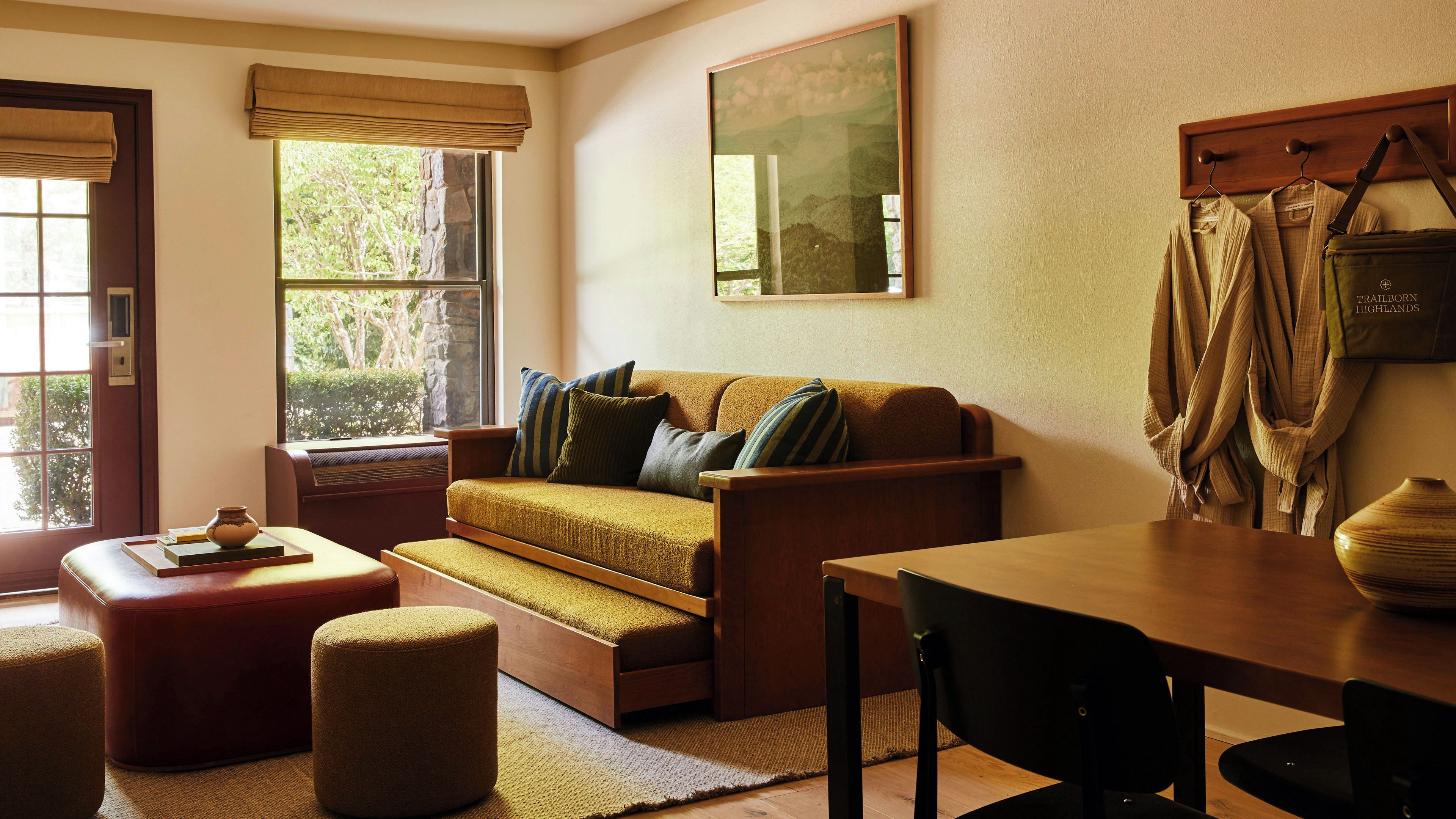 A cozy living room with a sofa, ottoman, and a table. There is a window with a view of trees, a painting on the wall, and a coat rack with hanging clothes.