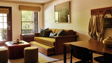Una sala de estar acogedora con un sofá, un reposapiés y una mesa. Hay una ventana con vistas a árboles, un cuadro en la pared y un perchero con ropa colgada.