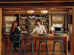 A bar area with wooden shelves stocked with bottles, a bartender, and two bar stools. - Highlands Inn Lodge Formerly Hampton Inn Highlands (Highlands)