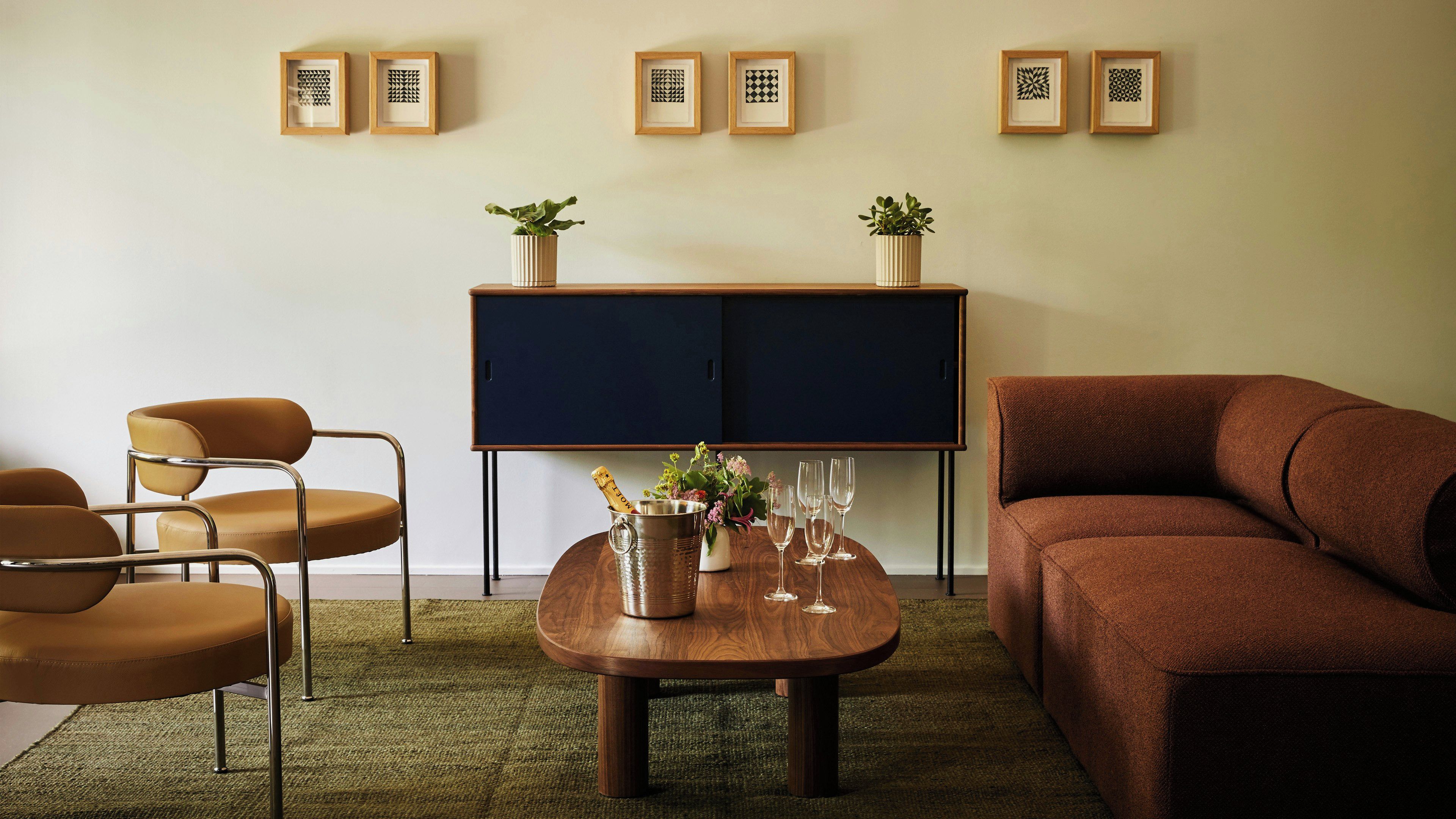 A living room with a brown sofa, a wooden coffee table with a bottle and glasses, a blue cabinet with potted plants, and a chair with a metal frame.