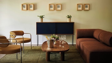 Una sala de estar con un sofá marrón, una mesa de centro de madera con una botella y vasos, un mueble azul con plantas en macetas, y una silla con estructura metálica.