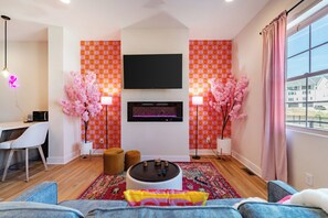 A modern living room with a flat-screen TV, two pink artificial cherry blossom trees, a dining area with a white chair, and a large window with a view of buildings. - Double Feature - Group Stay w/ Rooftop Hot tubs (Nashville)