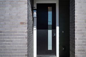 A modern door with a vertical glass panel, a black exterior, and a small outdoor light fixture on the wall. - Double Feature - Group Stay w/ Rooftop Hot tubs (Nashville)