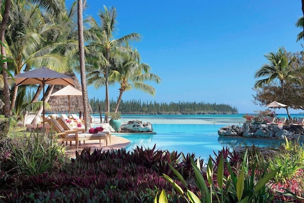 Un espace piscine agrémenté de chaises longues et de parasols, entouré de palmiers et de plantes tropicales.