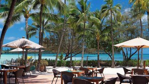 An outdoor dining area with tables and chairs, surrounded by palm trees and overlooking a beach. - Le Meridien Ile des Pins (L'Île-des-Pins)