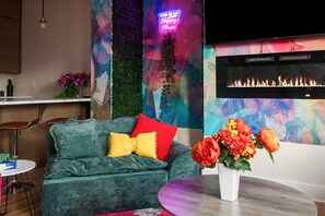 A cozy living area with a green sofa, a red and yellow pillow, a small round table with a vase of flowers, and a fireplace with a colorful abstract mural behind it.