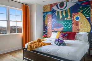 Ein Schlafzimmer mit einem großen Bett, einem Fenster mit Vorhängen und einer farbenfrohen abstrakten Wandkunst.