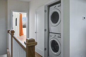Ein Waschraum mit zwei übereinandergestapelten Waschmaschinen, einer Holztreppe und einer Tür zu einem Raum mit einem orangefarbenen Vorhang.