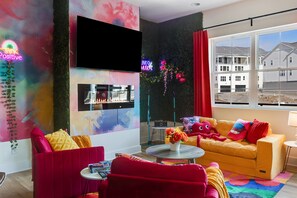 A modern living room with a flat-screen TV, a fireplace, colorful wall art, and a large window with a view of residential buildings. - Neon Nights - Big Groups/Game Room/Rooftop Hot Tub (Nashville)