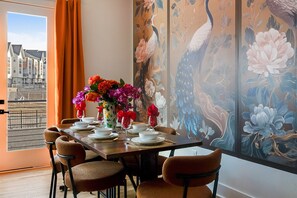 A dining area with a table set for four, a vase of flowers, and a large window with a view of buildings. - Velvet Escape - Groups/Game Room/Rooftop Hot Tub (Nashville)