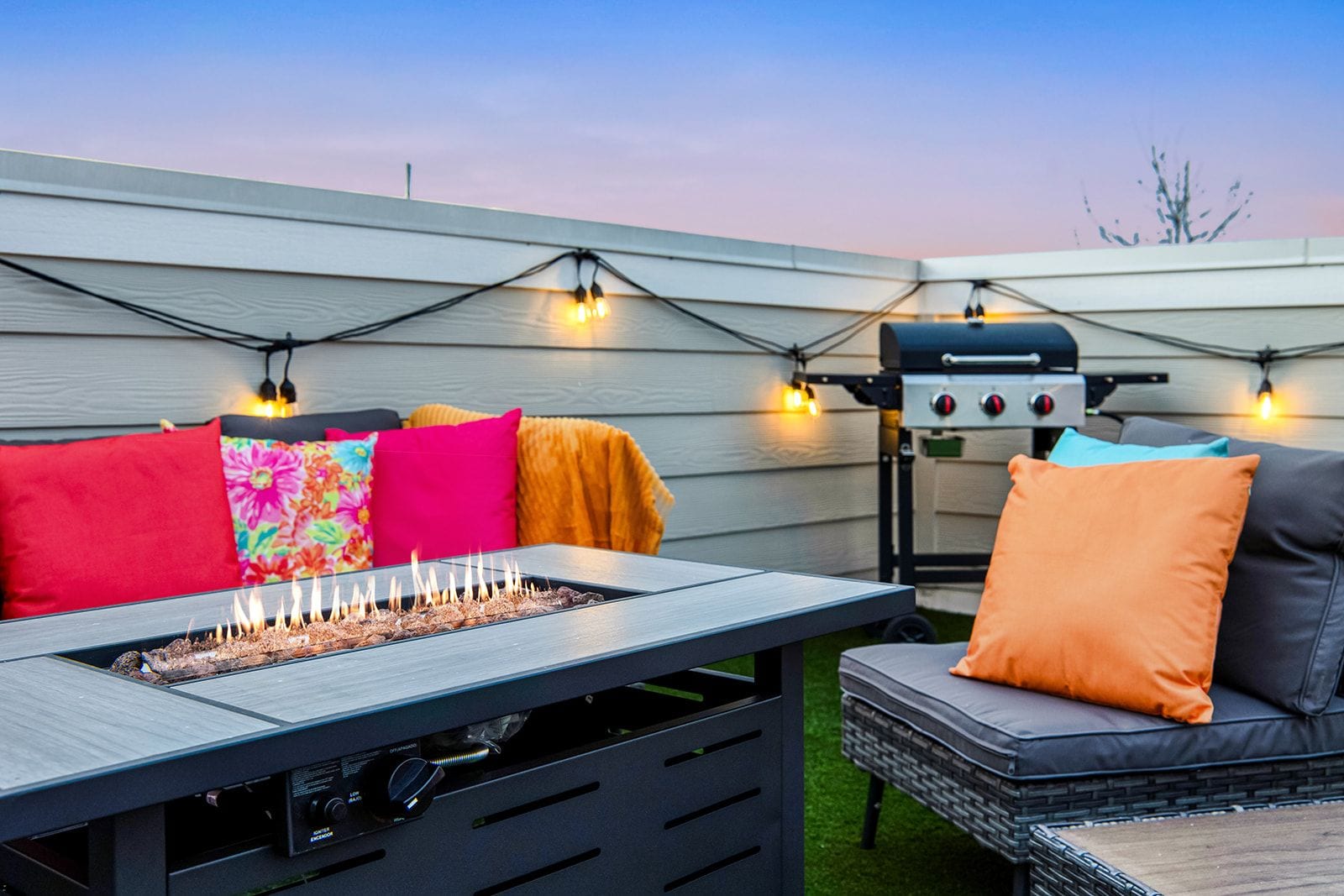 Eine Außenpatio mit Grill, einem Tisch mit Feuerstelle und farbenfrohen Kissen.