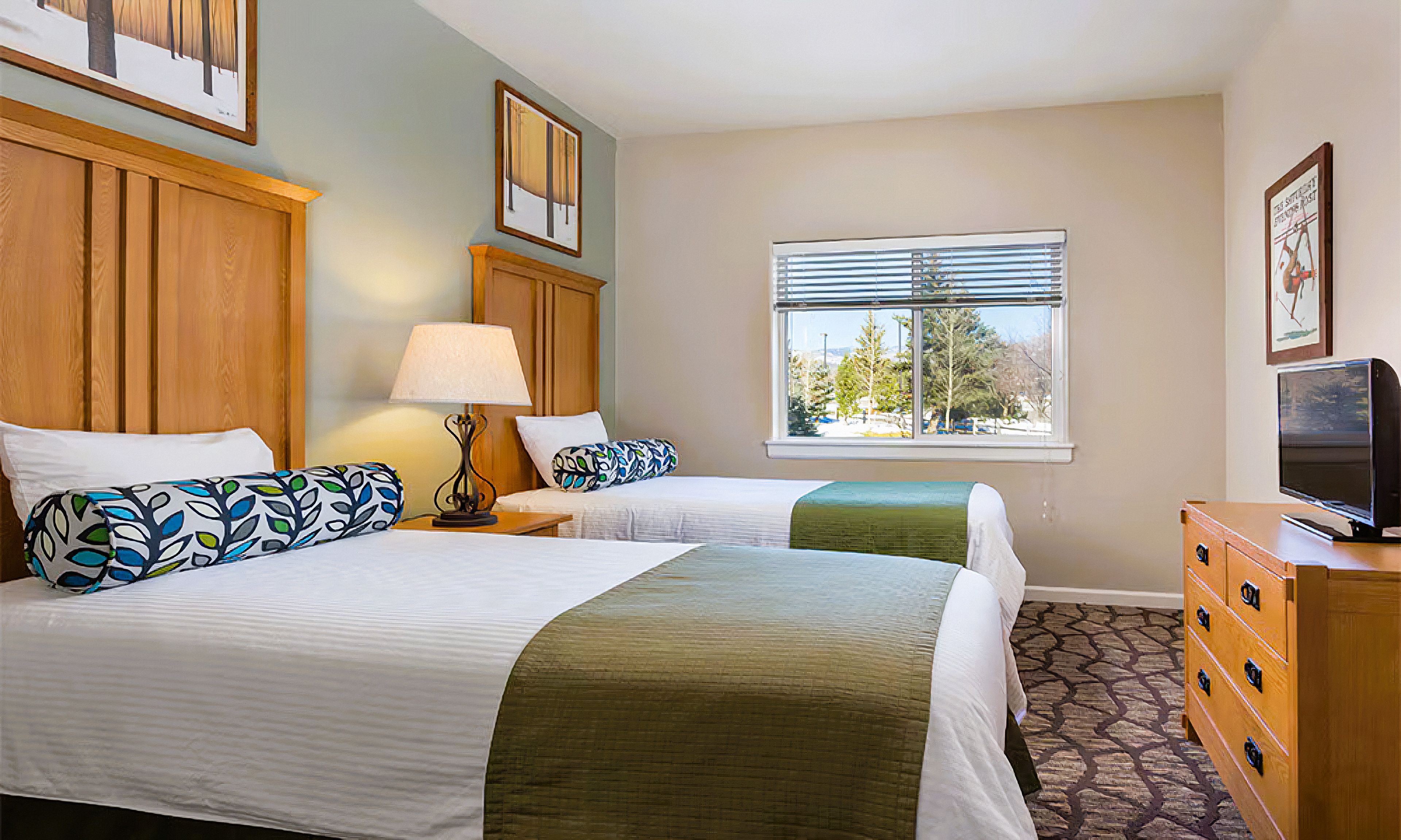 A hotel room with two beds, a dresser, a television, and a window with a view of trees.