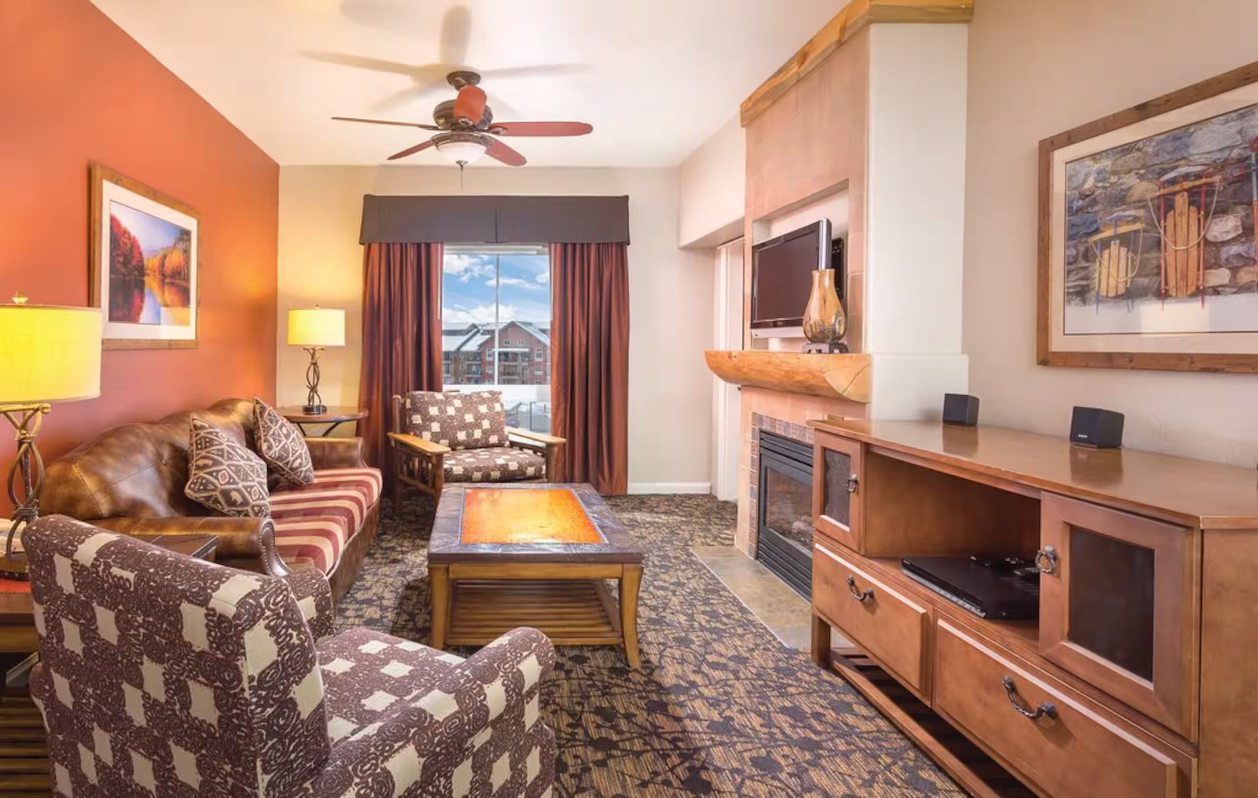 A hotel room with a sofa, armchair, coffee table, TV stand, fireplace, and a ceiling fan.