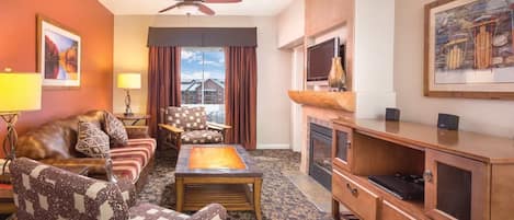 A hotel room with a sofa, armchair, coffee table, TV stand, fireplace, and a ceiling fan.