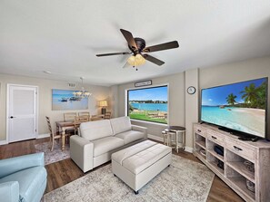 A living room with a white sofa, a television, a ceiling fan, a dining area with a table and chairs, and a large window with a view of a beach. - Compass Point 109 & Lagoon View (Gulf Shores)