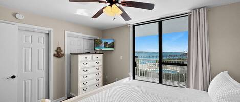 A bedroom with a bed, a ceiling fan, a dresser, a television, and a balcony with a view of the sea.
