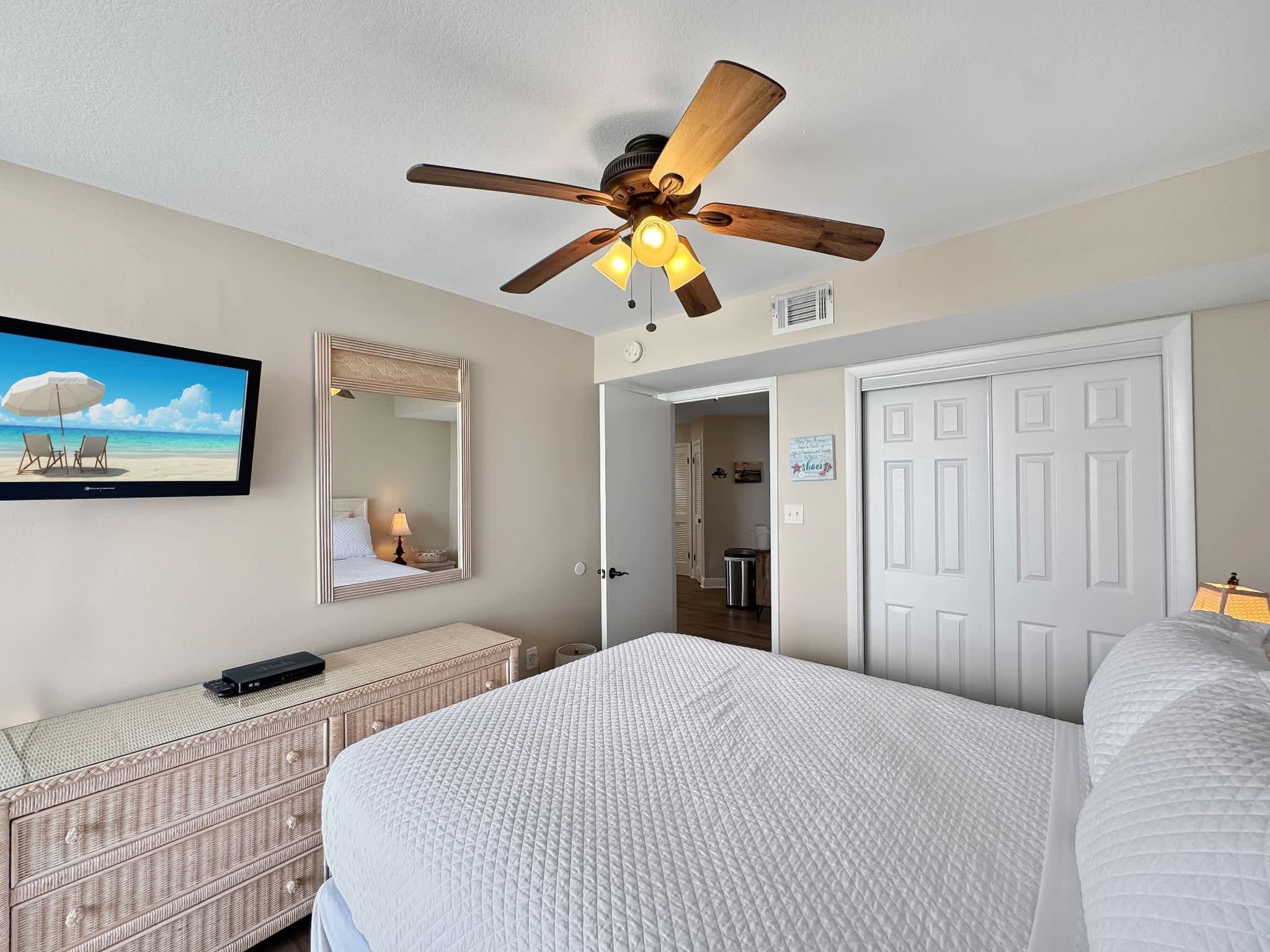 A bedroom with a bed, a ceiling fan, a television, a mirror, and a door leading to a bathroom.