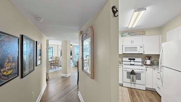 A hallway leading to a kitchen with white cabinets, a microwave, and an oven. There are framed pictures on the wall and a mirror.