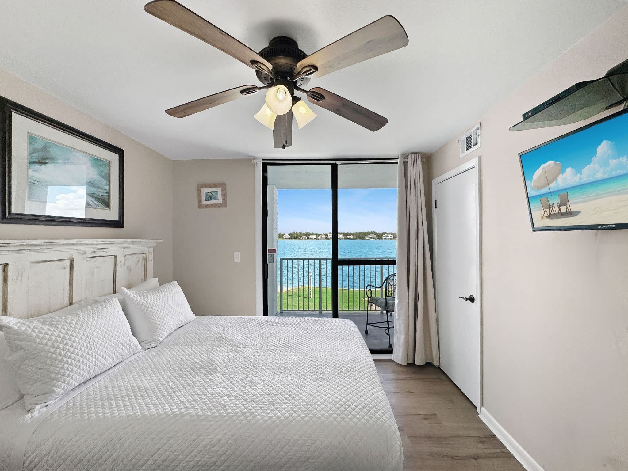 A bedroom with a bed, a ceiling fan, a view of the water through a glass door, and a painting on the wall.