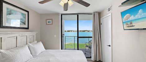 A bedroom with a bed, a ceiling fan, a view of the water through a glass door, and a painting on the wall.