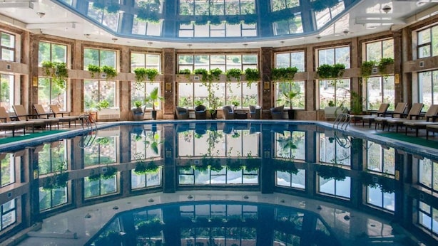 A large indoor swimming pool with a central island, surrounded by lounge chairs and potted plants. - Jannat Resort Koi-tash (Koy-Tash)