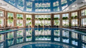 Una amplia piscina interior con paredes de vidrio transparente, rodeada de sillones de descanso y plantas en macetas.