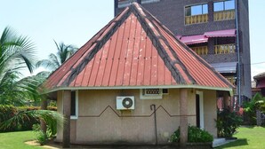 Un petit bâtiment d’un seul  étage, avec un toit rouge, entouré de verdure et avec un grand immeuble en arrière-plan.
