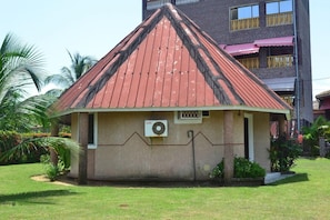 A small, single-story building with a red roof, surrounded by greenery and a tall building in the background. - Oceanic Beach Hotel (Kribi)