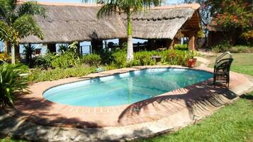 A swimming pool with a brick border, surrounded by grass and palm trees, with a thatched-roof structure in the background.