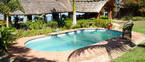 Une piscine bordée de briques, entourée de pelouse et de palmiers, avec une structure à toit de chaume en arrière-plan.