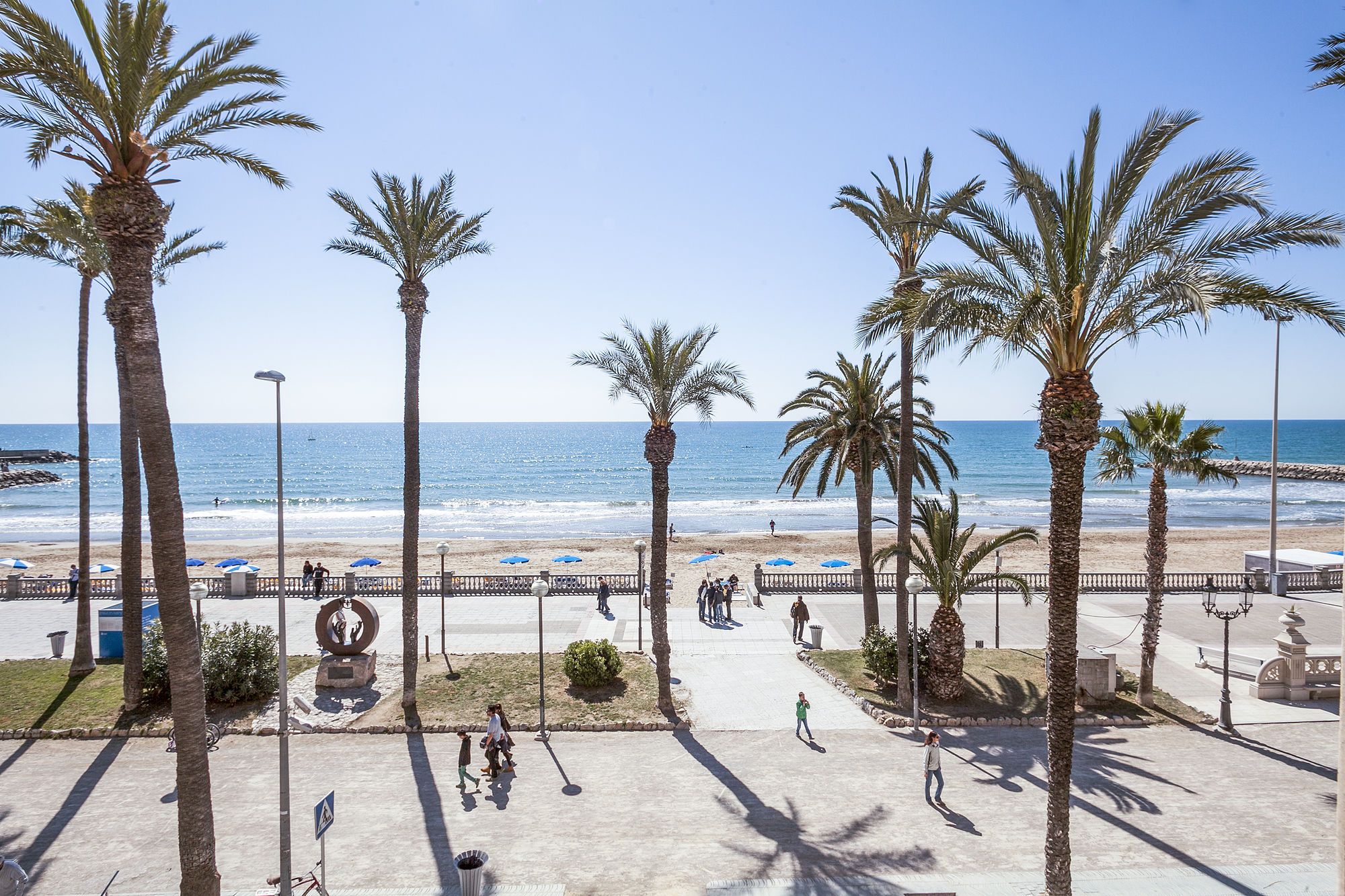 A coastal promenade with palm trees, a beach, and a clear sky.
