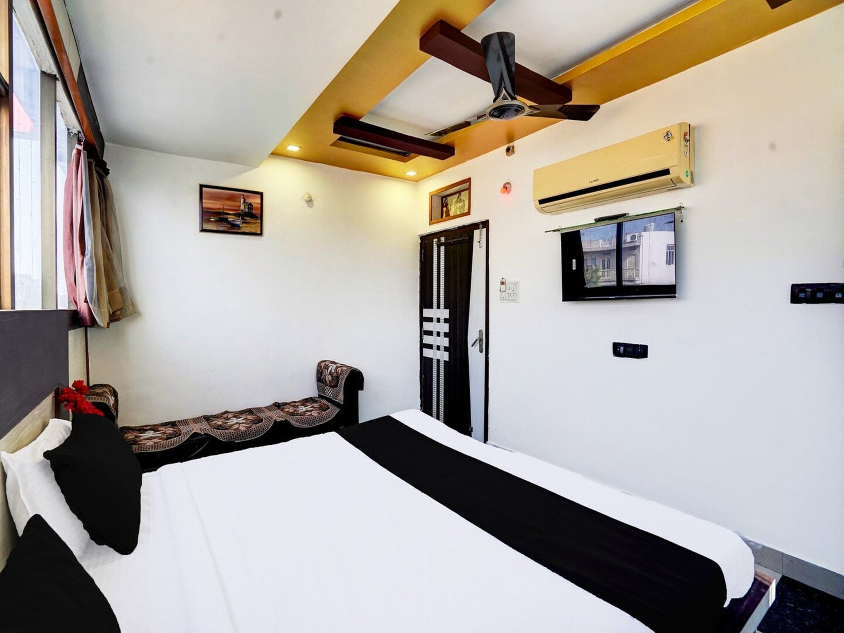 A hotel room with a bed, a ceiling fan, an air conditioning unit, a door with a decorative handle, and a window with curtains.