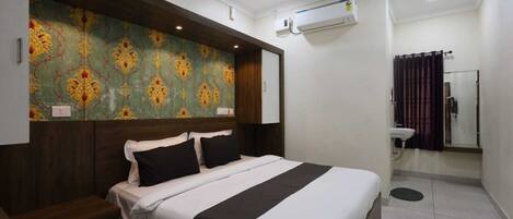 Habitación de hotel con cama, ventilador de techo, equipo de aire acondicionado y baño con lavamanos y espejo.