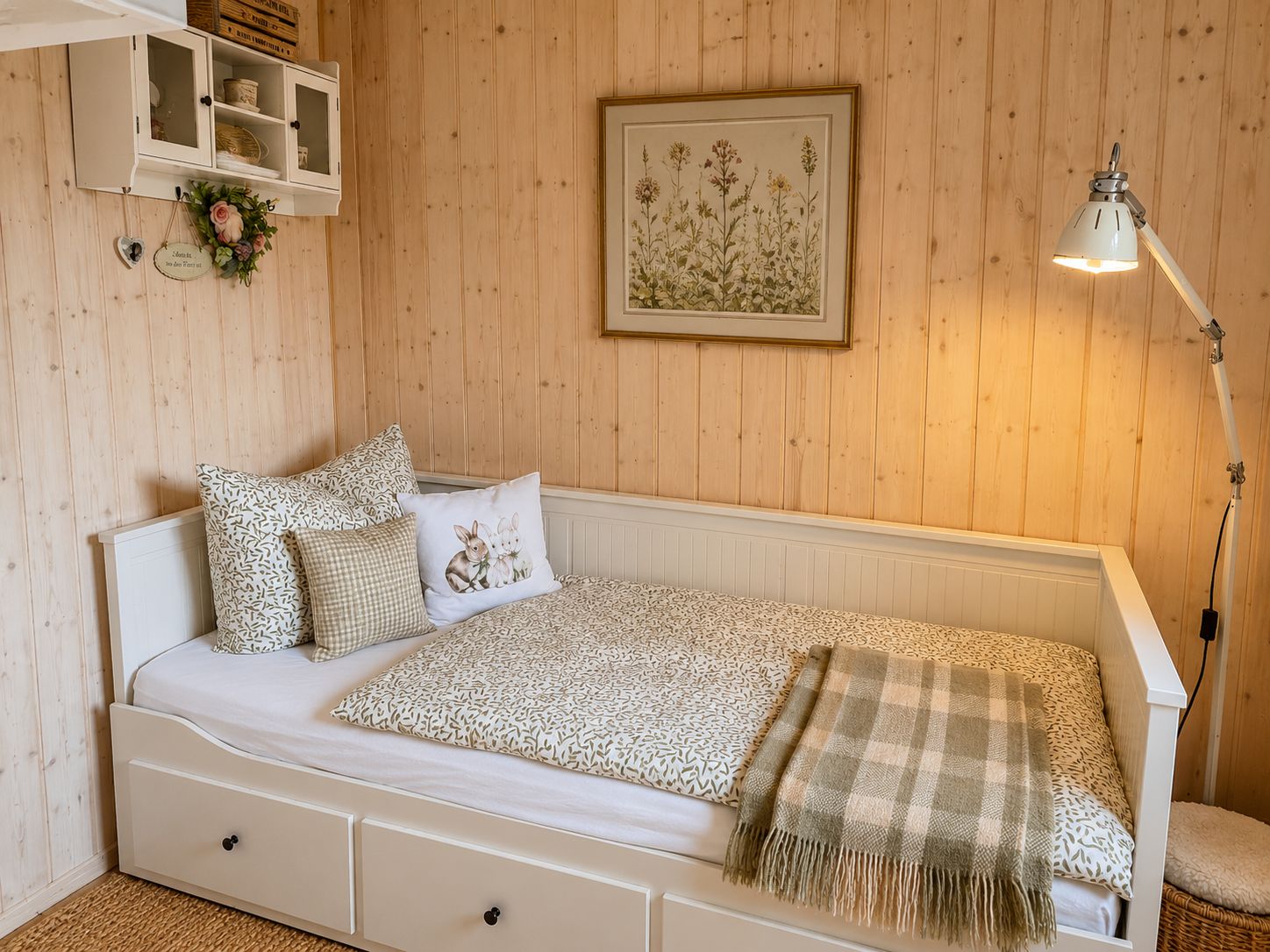 A single-bed room with a wooden wall, a framed botanical illustration, a bedside lamp, and a small shelf with decorative items.