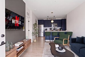 Ein modernes Wohnzimmer mit einem Flachbildfernseher, einer Essecke mit Tisch und Stühlen sowie einer Küche mit dunklen Schränken und einem Edelstahlkühlschrank.