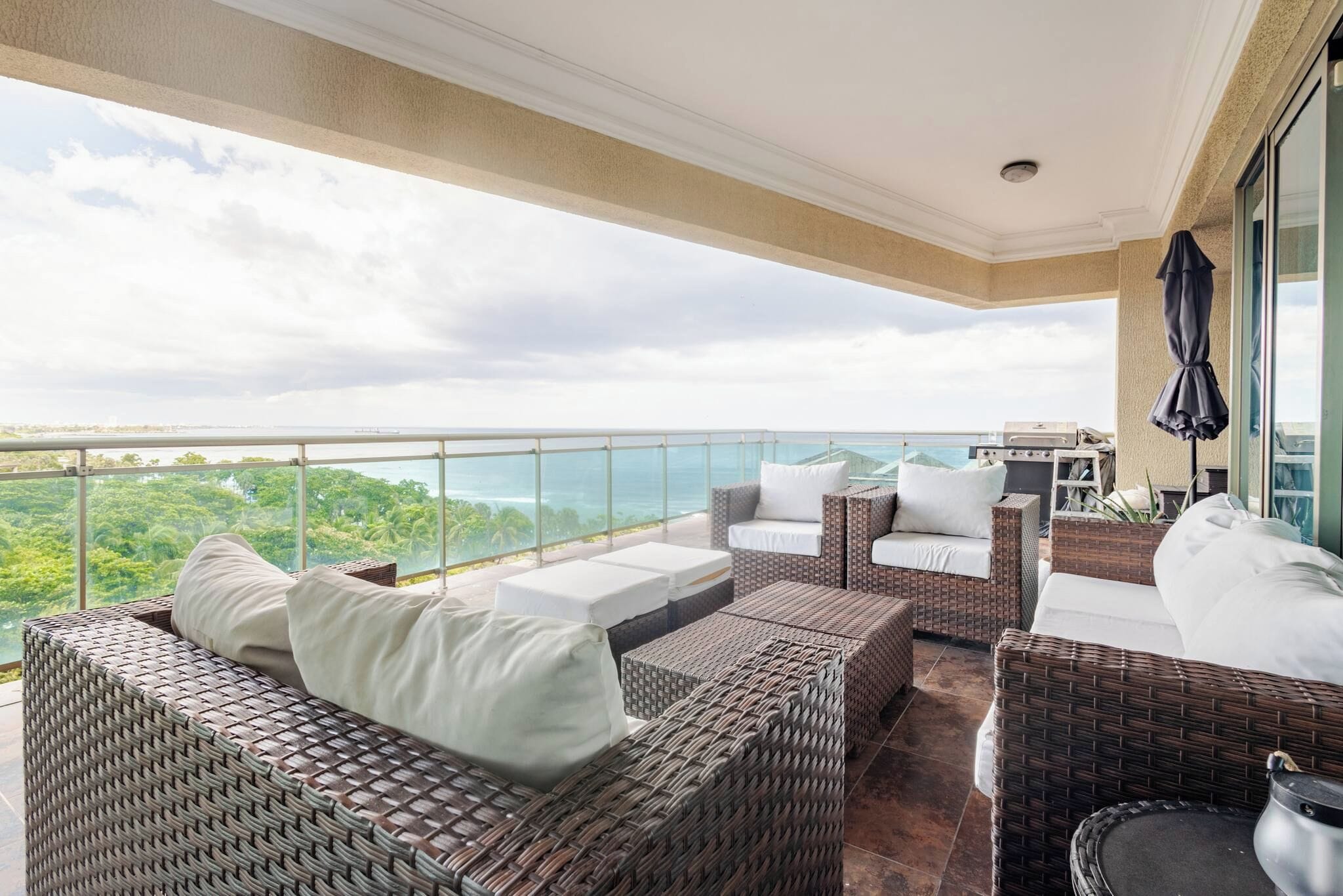 A balcony with wicker furniture, a grill, and a view of the ocean.