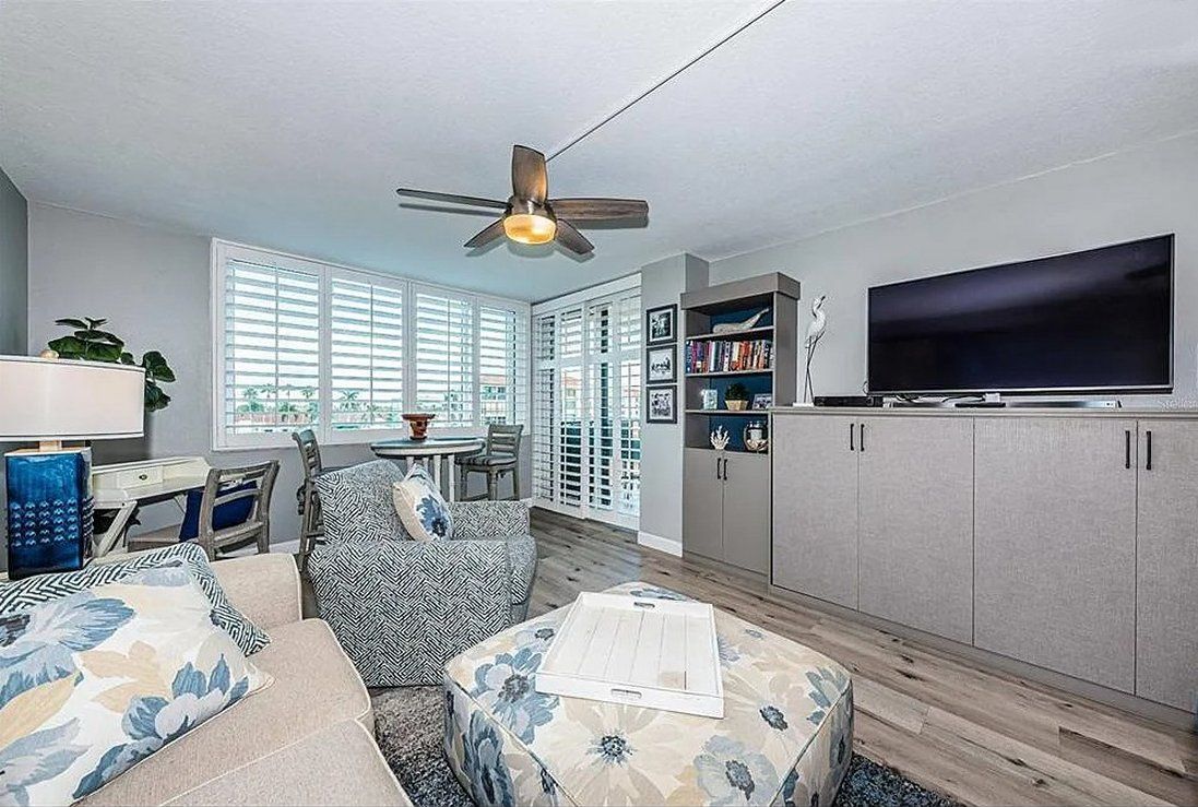A living room with a sofa, ottoman, ceiling fan, TV, and kitchen area with cabinets.