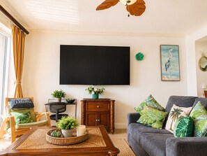 Ein Wohnzimmer mit an der Wand montiertem Flachbildfernseher, einem Sofa mit tropischen Kissen, einem Couchtisch aus Holz mit Pflanze und Kerze sowie einem Sessel mit einem 'Welcome'-Schild.
