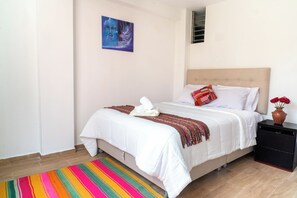 Ein ordentlich bezogenes Bett mit weißen Leinen, ein farbenfroher Streifen Teppich, ein kleiner Nachttisch mit Blumenvase und ein Gemälde an der Wand.