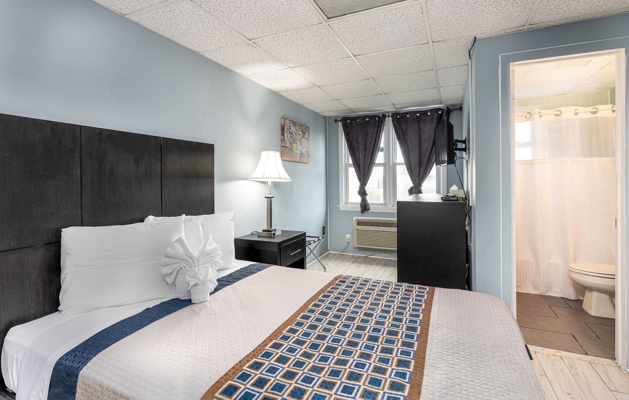 A hotel room with a bed, a nightstand, a lamp, a window with curtains, and a bathroom visible through an open door.